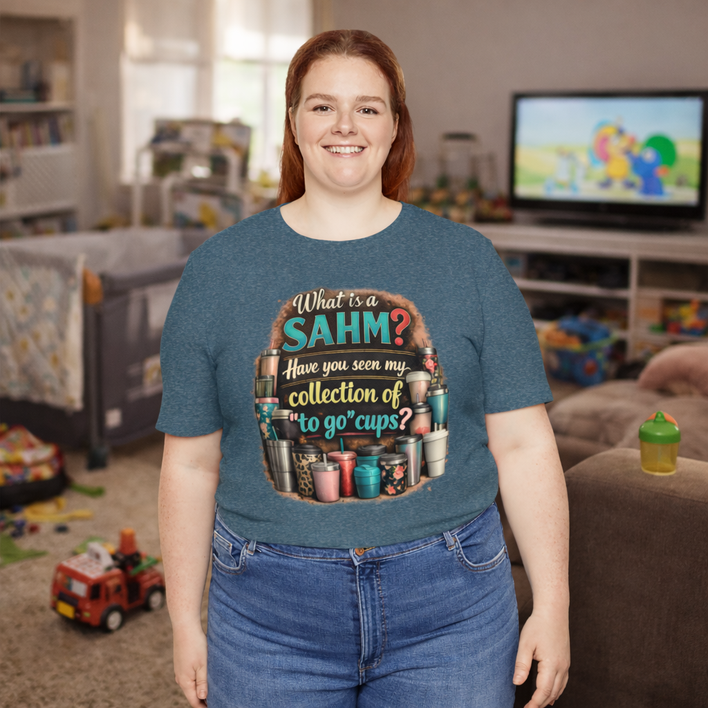 Smiling stay at home mom wearing a teal SAHM coffee graphic t-shirt with a humorous quote about collecting to-go cups, standing in a cozy living room with kids toys in the background.