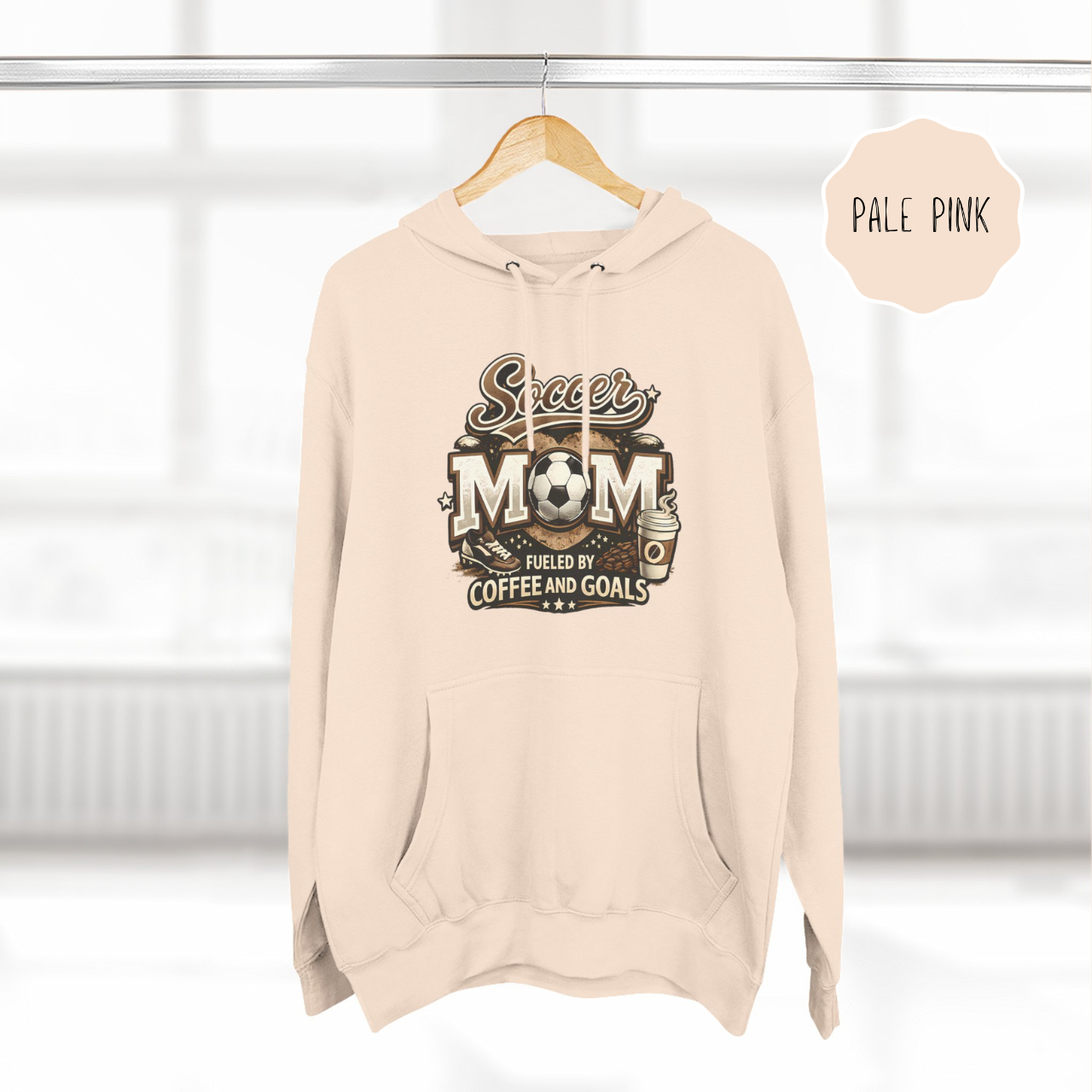 Pale Pink color soccer mom hoodie hanging on hanger with coffee and goals design
