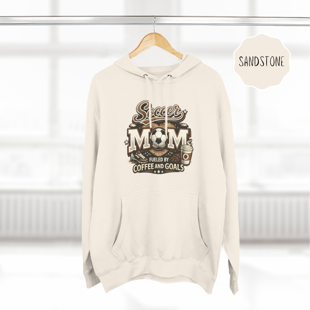 Neutral color soccer mom hoodie hanging on hanger with coffee and goals design