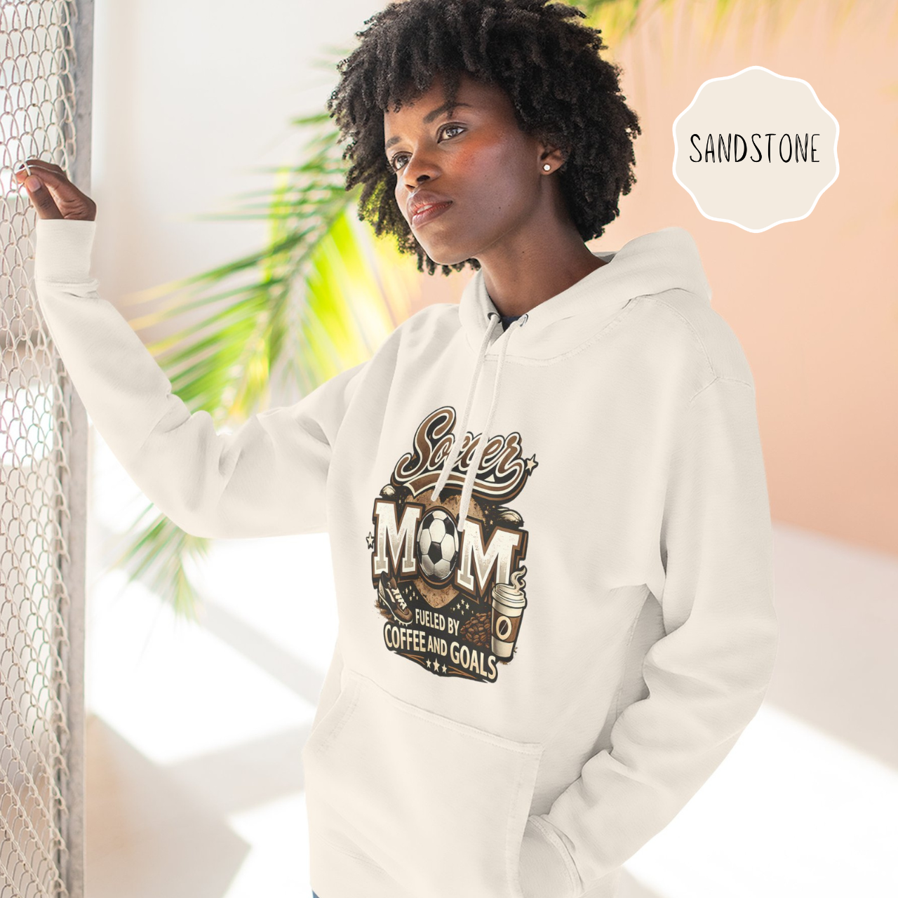 Woman wearing a light color soccer mom hoodie with coffee and goals graphic outdoors